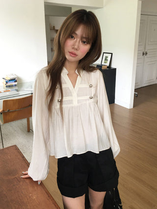 Model wearing a taupe gray long-sleeve pleated blouse with front pockets and rounded button accents